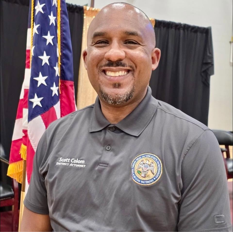 Scott Colom smiling in a District Attorney polo shirt, with an American flag behind him